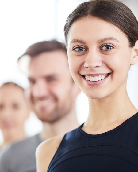 Smiling attractive teenage gymnast with pretty smile standing in row of professionals and looking at camera